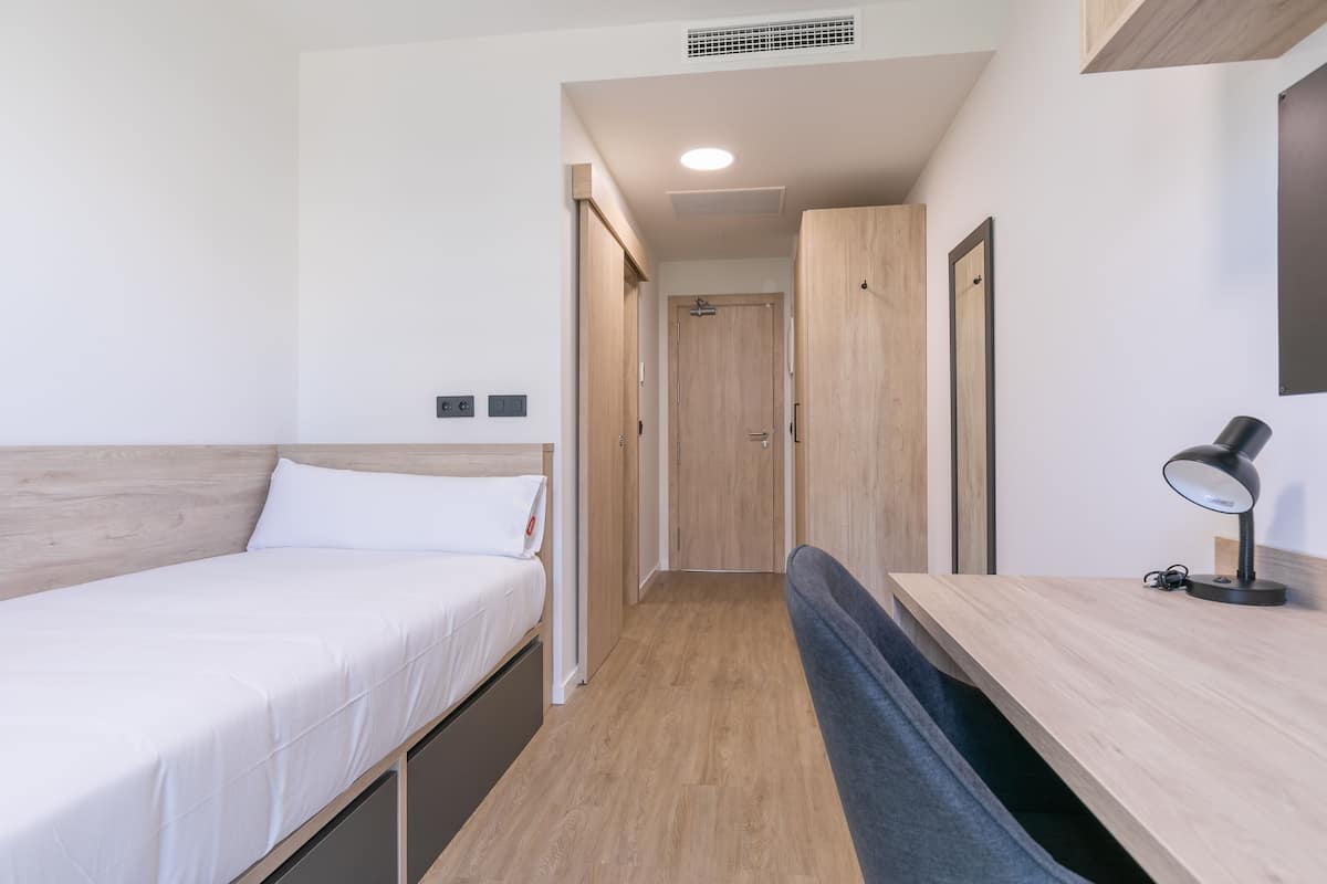 Una habitación pequeña con una cama, un escritorio y una silla. Hay una puerta que da a otra habitación.