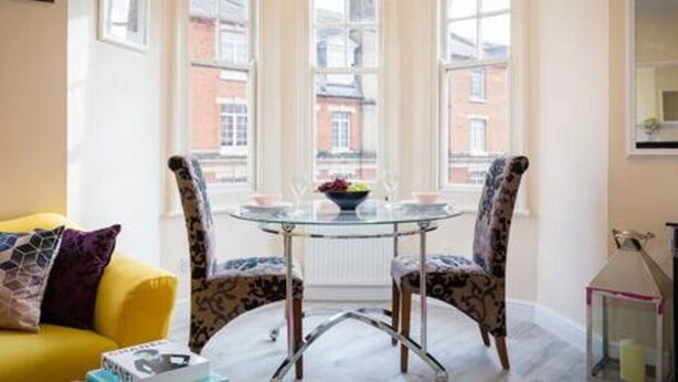 Una habitación con una mesa de vidrio, dos sillas estampadas, un sofá amarillo y vistas a edificios a través de amplias ventanas.