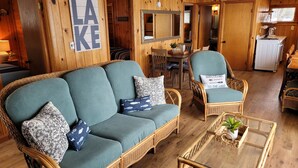Ein gemütliches Blockhaus-Interieur mit einem grünen Sofa, Korbstühlen, einem Esstisch und einer Küchenzeile.