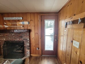 A wooden interior with a brick fireplace, a sign reading "WINNIPESAUKEE", and a door leading outside. - Winni Lakefront Cabin (Laconia)