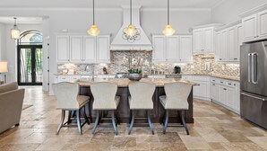 Una cocina amplia con muebles blancos, una isla central con taburetes de bar, un refrigerador de acero inoxidable e iluminación colgante.