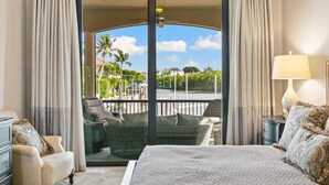 Habitación de hotel con una cama grande, un sofá, una silla, una mesita de noche y vistas a un balcón con mobiliario exterior y una masa de agua.