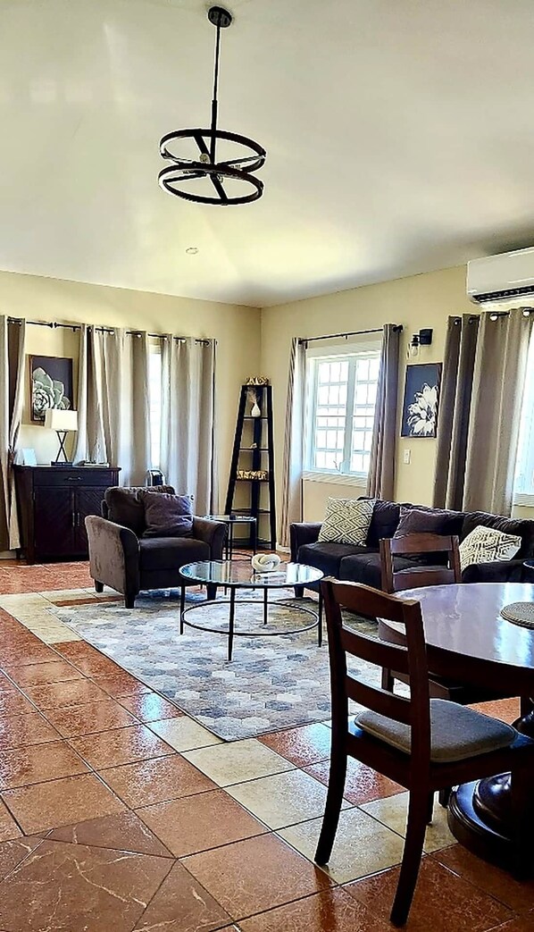 A living room with a sofa set, a coffee table, and a dining table with chairs. There is a ladder against the wall and a window with curtains.