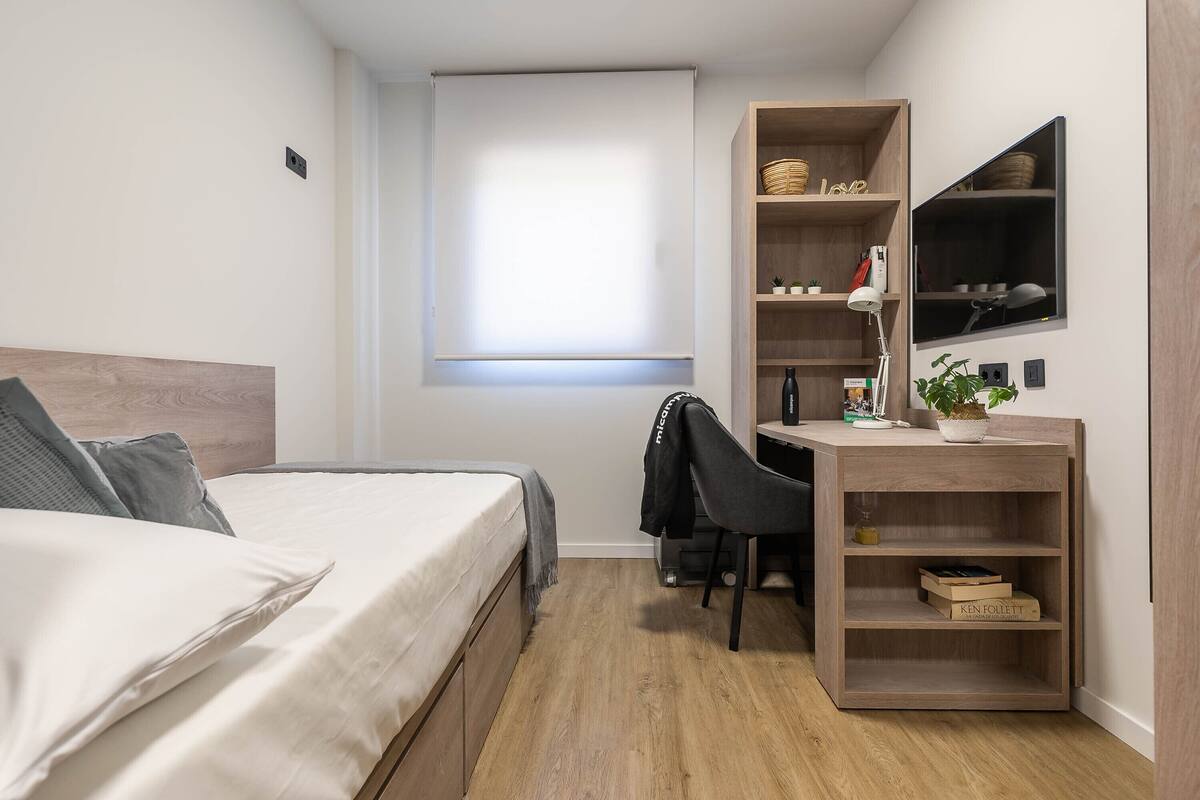 Un dormitorio con cama, escritorio, silla, estante con objetos y ventana.