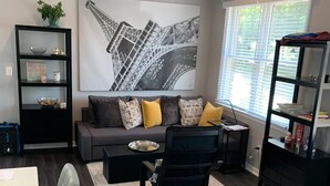 A modern living room with a grey sofa, black armchair, and a black bookshelf. There's a large black and white artwork on the wall, a window with blinds, and a dining area with a table and chairs. - Your Bright, Confortable, Cozy Second Home

 (Livingston)