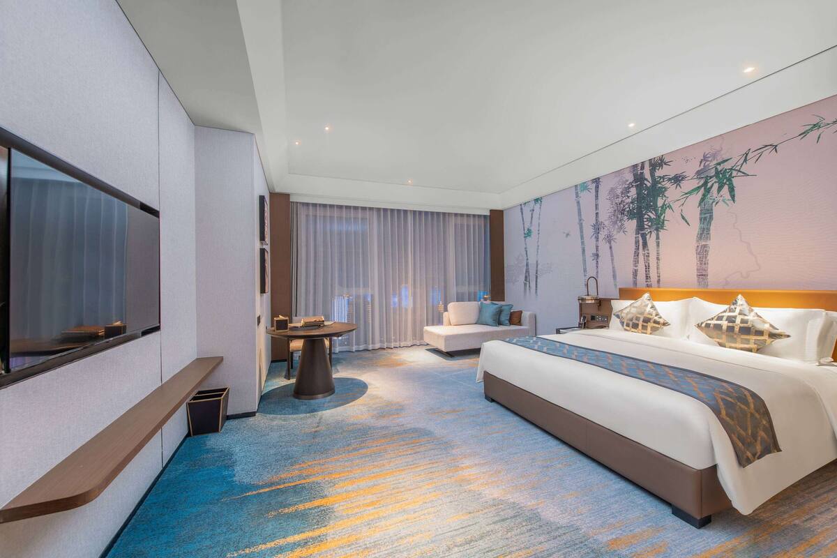 Ein modernes Hotelzimmer mit einem großen Bett, einer Sitzecke, einem Schreibtisch und einem großen Fenster mit Vorhängen.