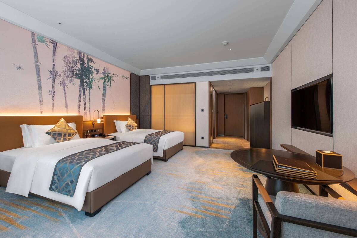 Ein Hotelzimmer mit zwei Betten, einem Fernseher, einem Schreibtisch und einer Wand mit einem Bambusgemälde.
