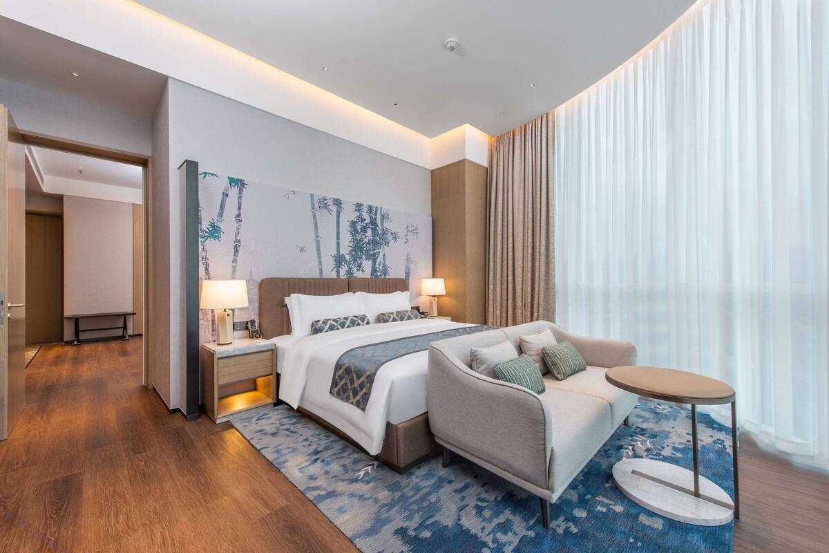 Ein modernes Hotelzimmer mit einem großen Bett, einer Couch, einem kleinen runden Tisch und einem großen Fenster mit Vorhängen.