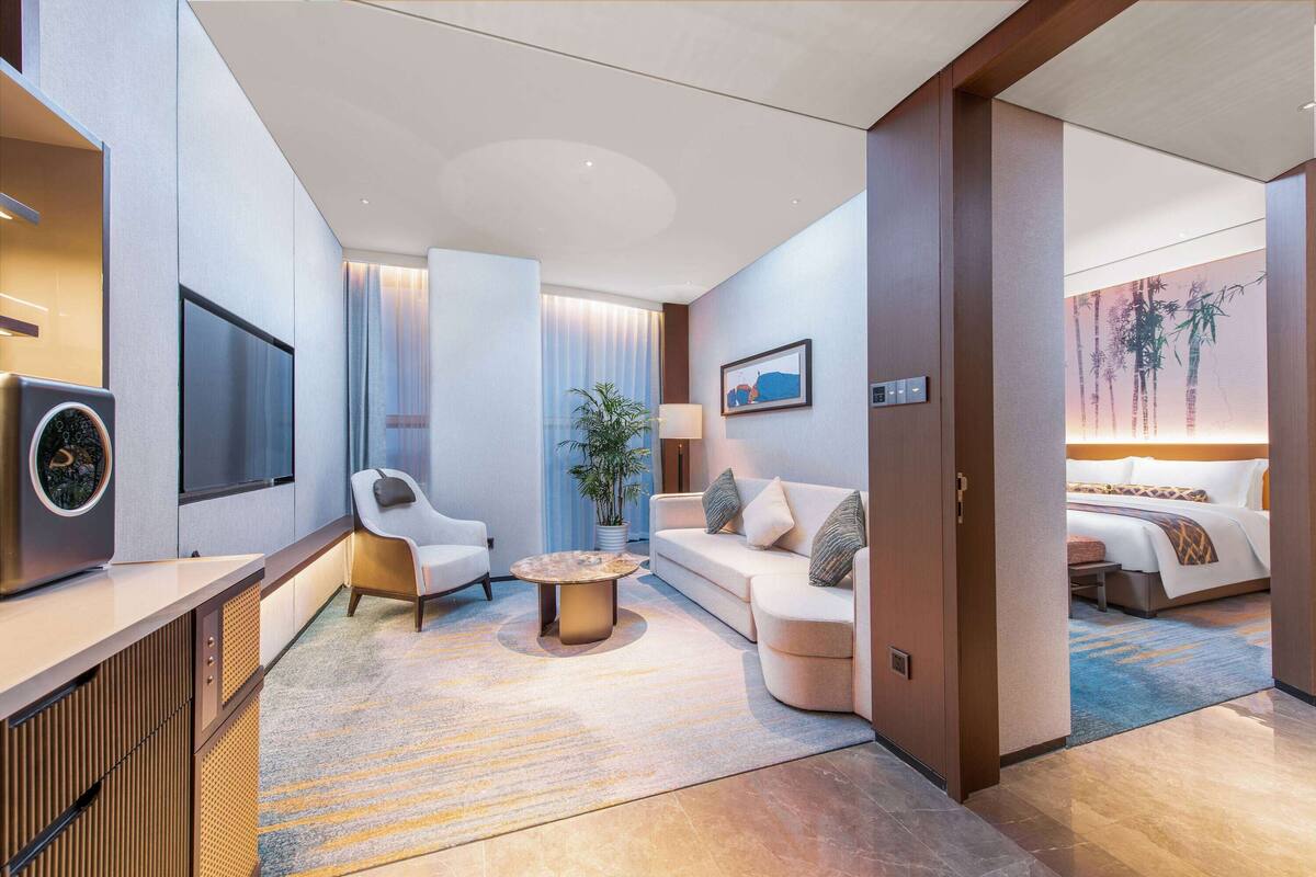 Ein modernes Hotelzimmer mit einer Couch, einem Sessel und einem kleinen Couchtisch. Ein Fernseher ist an der Wand montiert und ein Bett mit einem blau-braunen Musterdecke.
