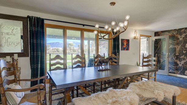 Une salle à manger avec une grande table, des chaises en bois, un lustre et une vue sur l’extérieur.