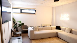 A modern living room with a sectional sofa, a small table with a laptop and remote control, a potted plant, and a wall-mounted projector displaying "Lugo House". - Micampus Lugo (Lugo)