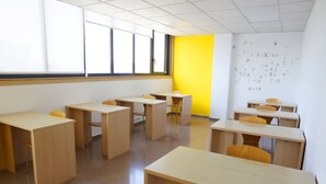 A classroom with wooden desks and chairs, large windows, and a yellow accent wall with alphabet letters. - Micampus Lugo (Lugo)