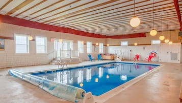 An indoor swimming pool with a clear blue water, surrounded by red and blue chairs, and a wooden ceiling with hanging lights.