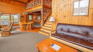 Ein Holzhütten-Interieur mit einer braunen Ledersofa, einem Holzesstisch und einer Treppe, die zu einem Schlafpodest führt.