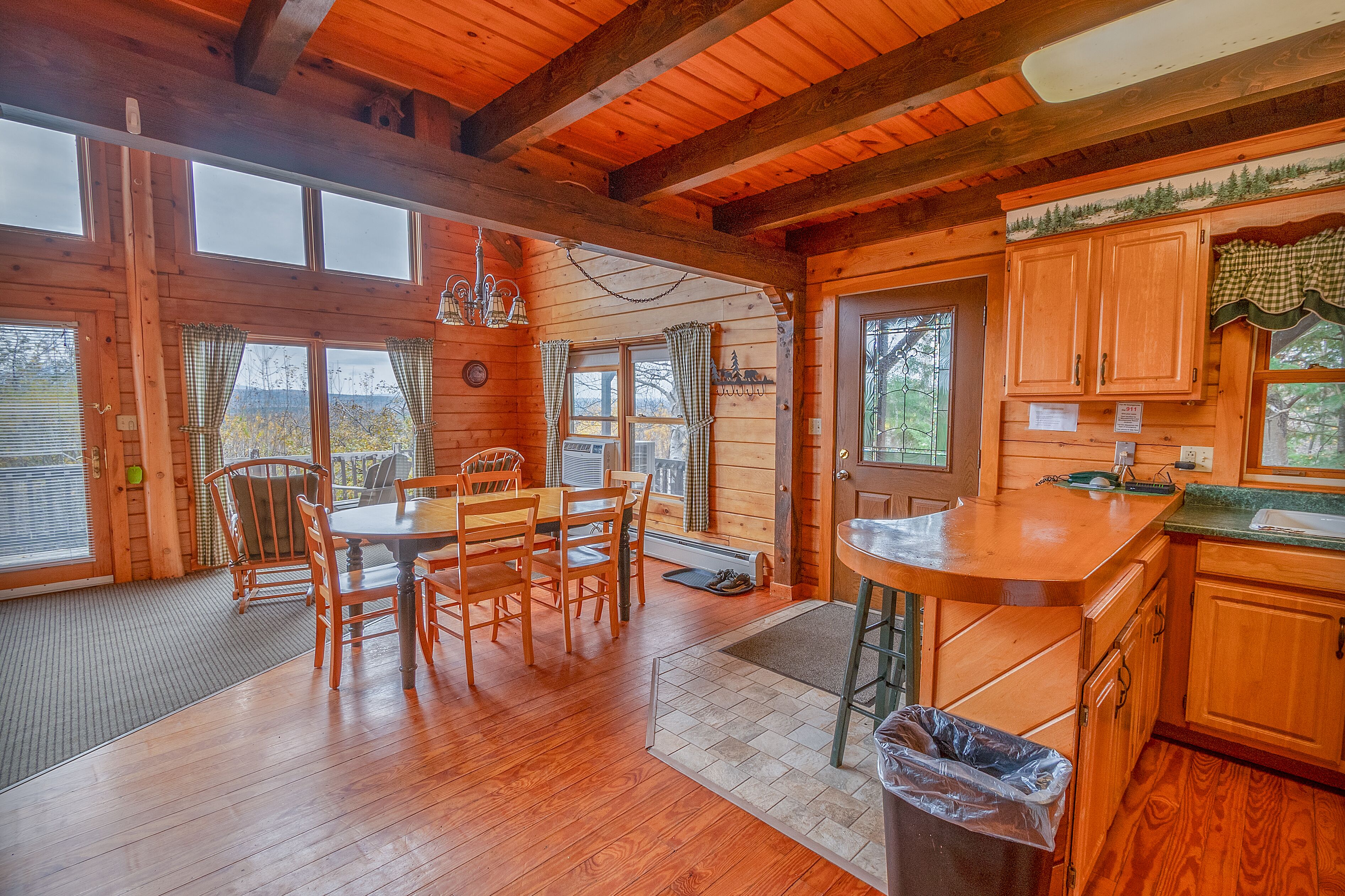 A wooden cabin interior with a dining area, kitchen, and large windows.
