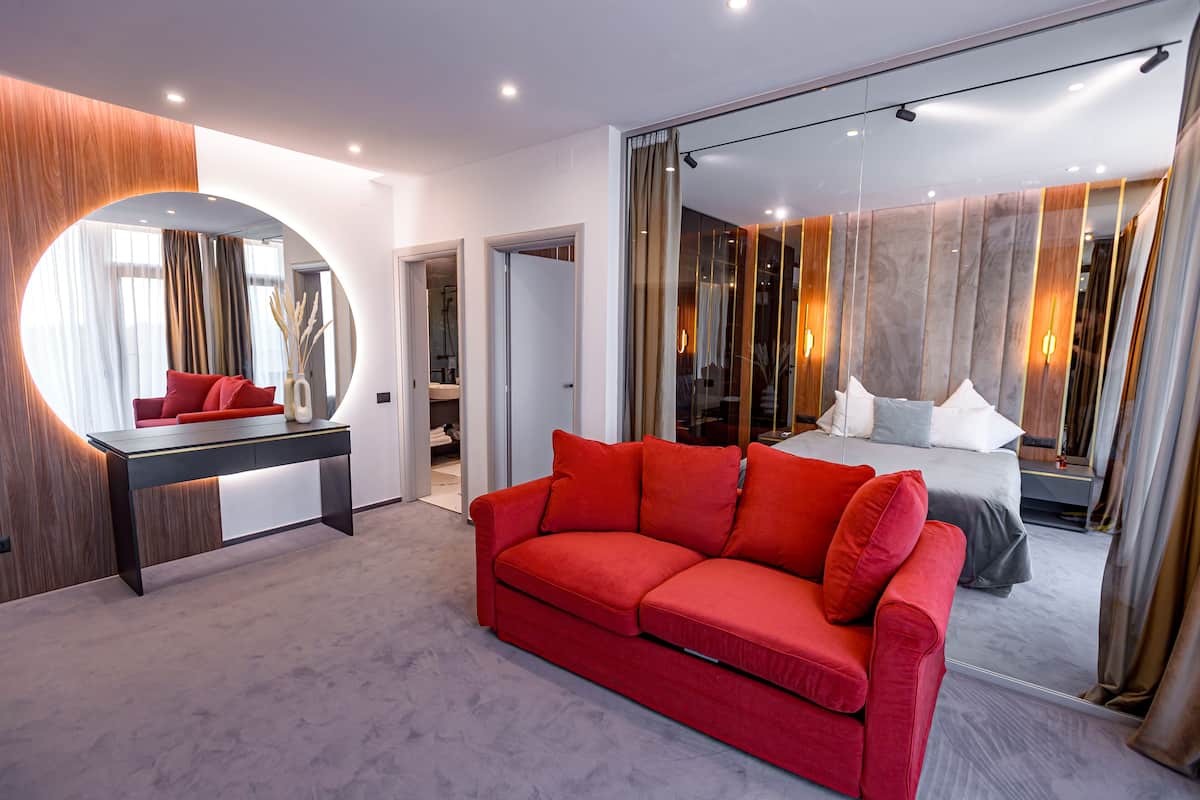 A modern hotel room with a large mirror, a red sofa, a bed with white and gray bedding, and a large window with curtains.