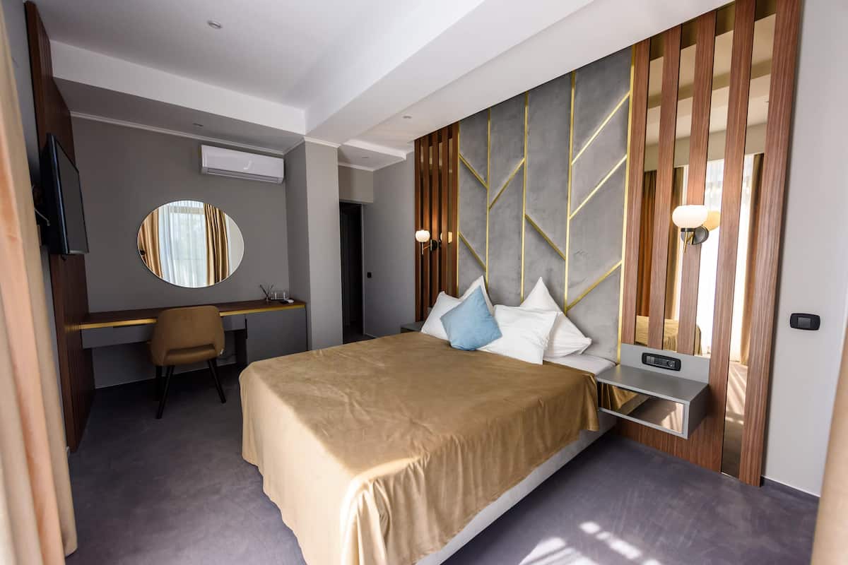 A modern hotel room with a bed, a desk, a chair, a TV, a round mirror, and a wall-mounted light fixture.