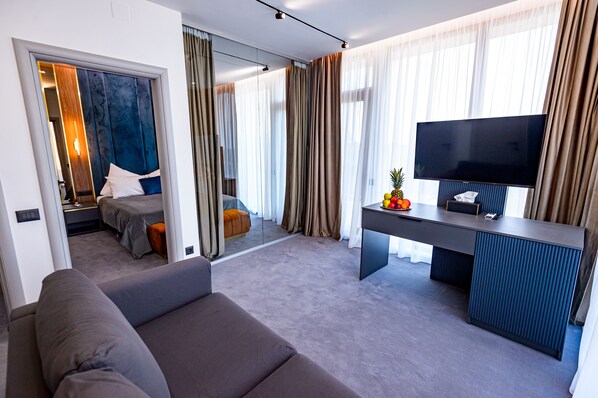 A modern hotel room with a large bed, a sofa, a desk with a flat-screen TV, and a large window with curtains.