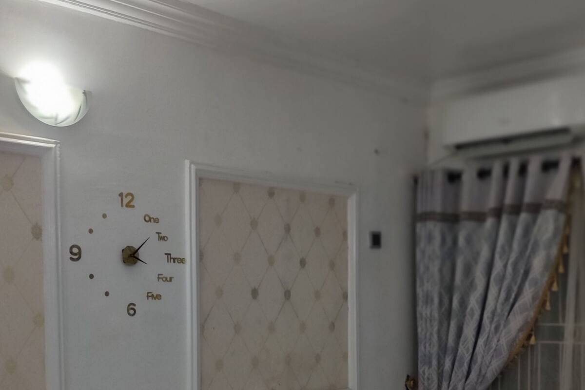 A room with a white sofa set, a wall clock, and a window with curtains.