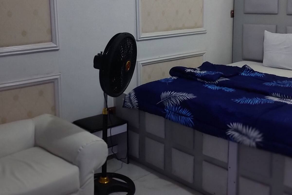 A bedroom with a bed, a chair, a standing fan, and a door.