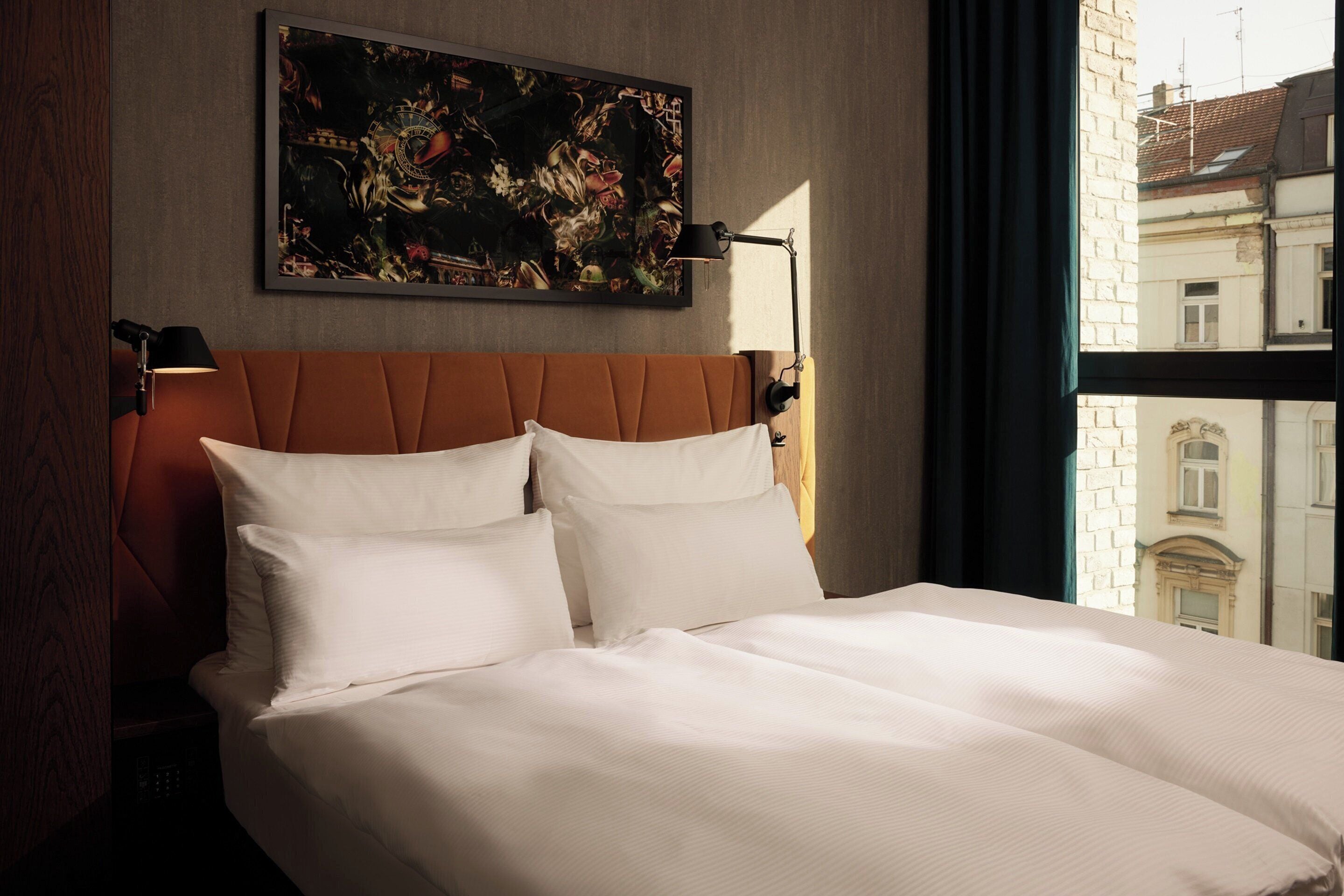A hotel room with a large bed, two bedside lamps, a painting on the wall, and a view of buildings through the window.