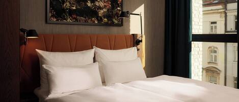 A hotel room with a large bed, two bedside lamps, a painting on the wall, and a view of buildings through the window.