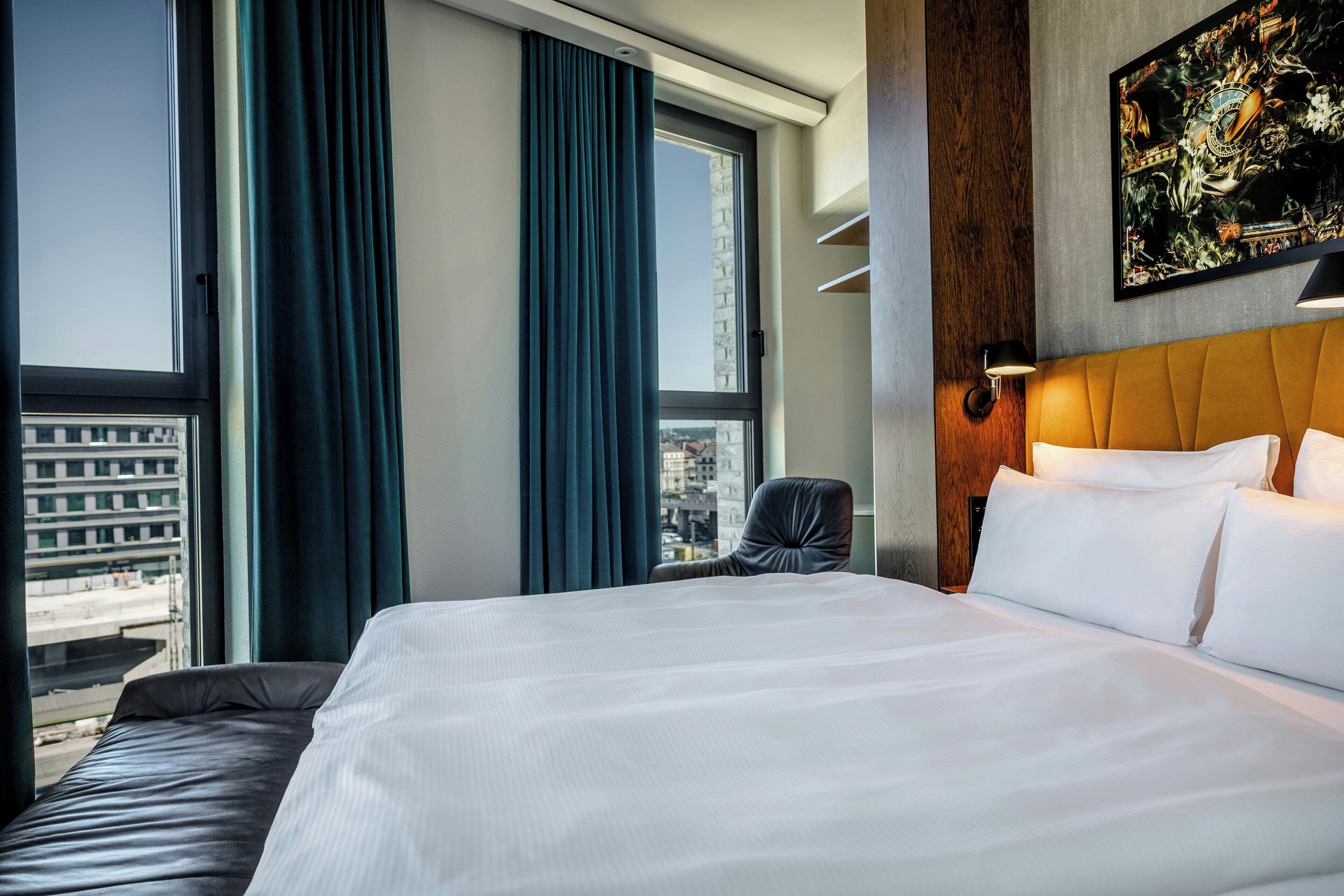Una habitación de hotel con una cama grande, una silla, una ventana con cortinas y una obra de arte colgada en la pared.
