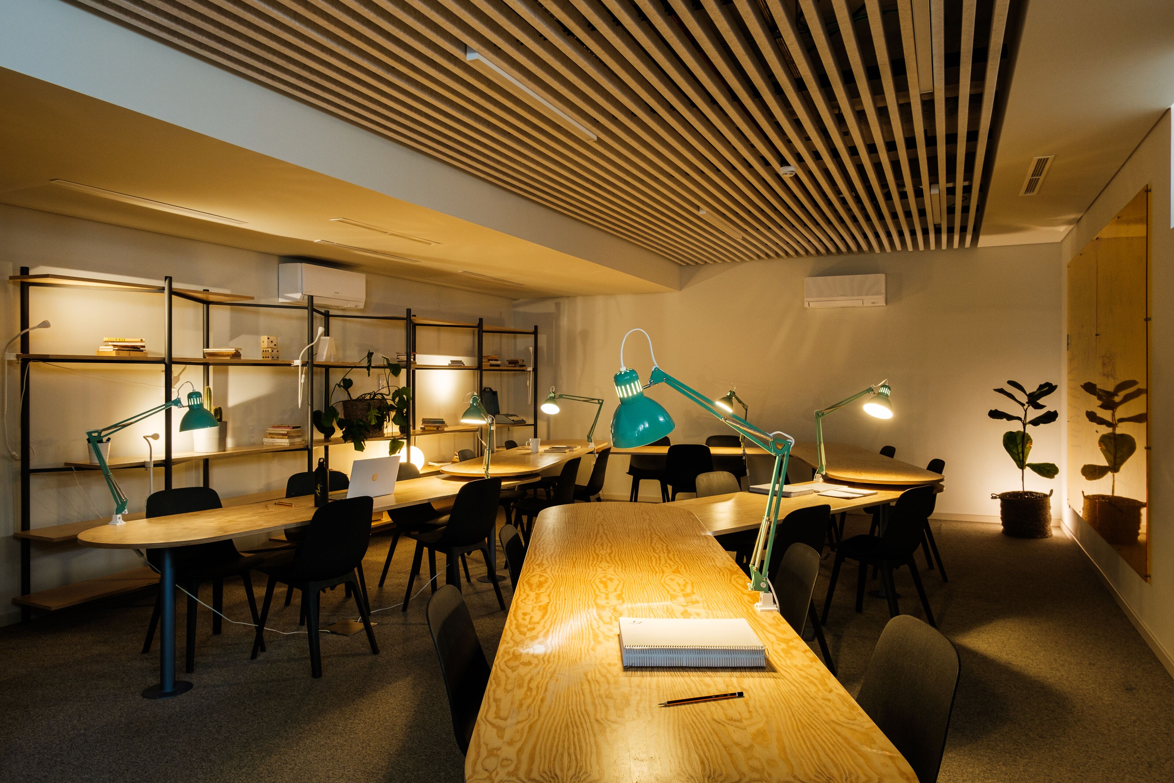 Una sala riunioni moderna con un lungo tavolo in legno, diverse sedie e lampade da tavolo verdi. Si notano scaffali con libri e piante sullo sfondo.