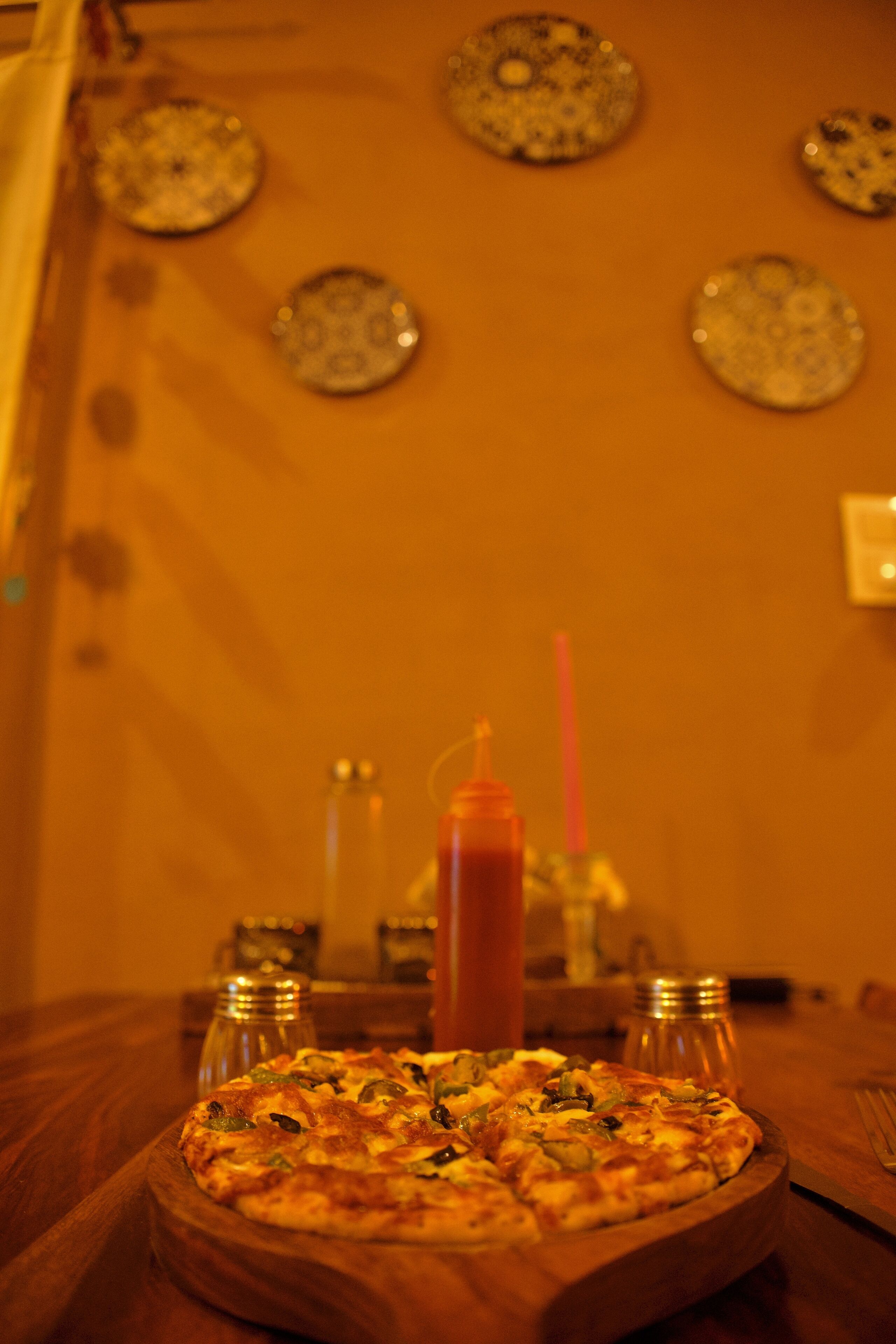 A pizza on a wooden board with a bottle of ketchup and two salt shakers on a table.
