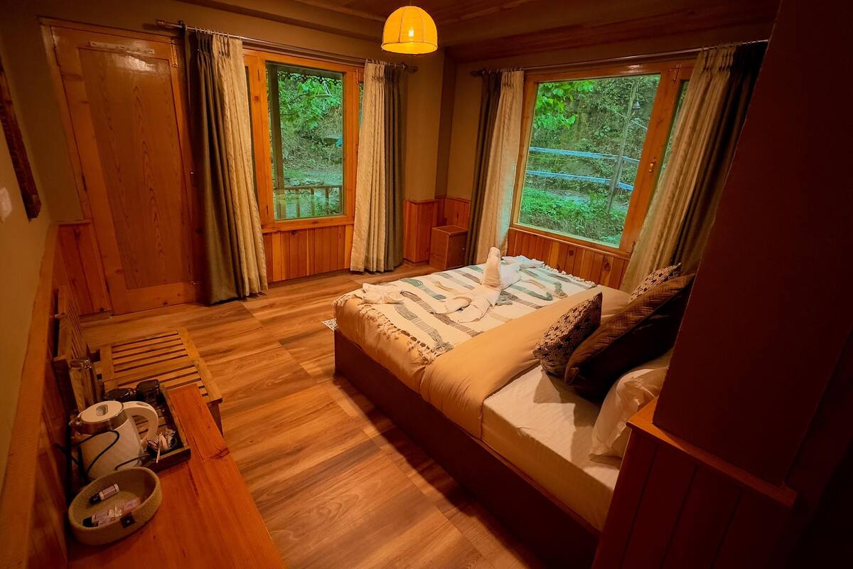 A cozy bedroom with a wooden bed, a bedside table, a window with curtains, and a view of greenery outside.