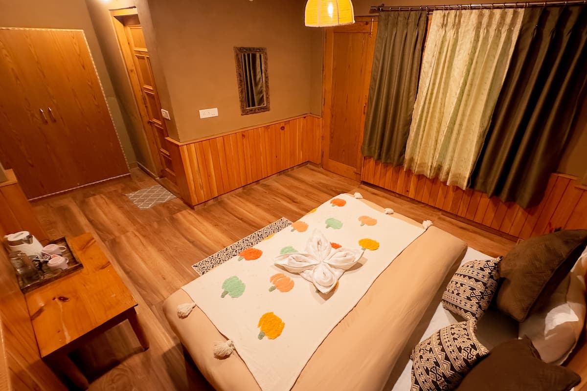 A bedroom with a wooden floor, a bed with a floral bedspread, a bedside table with a lamp, and a window with curtains.