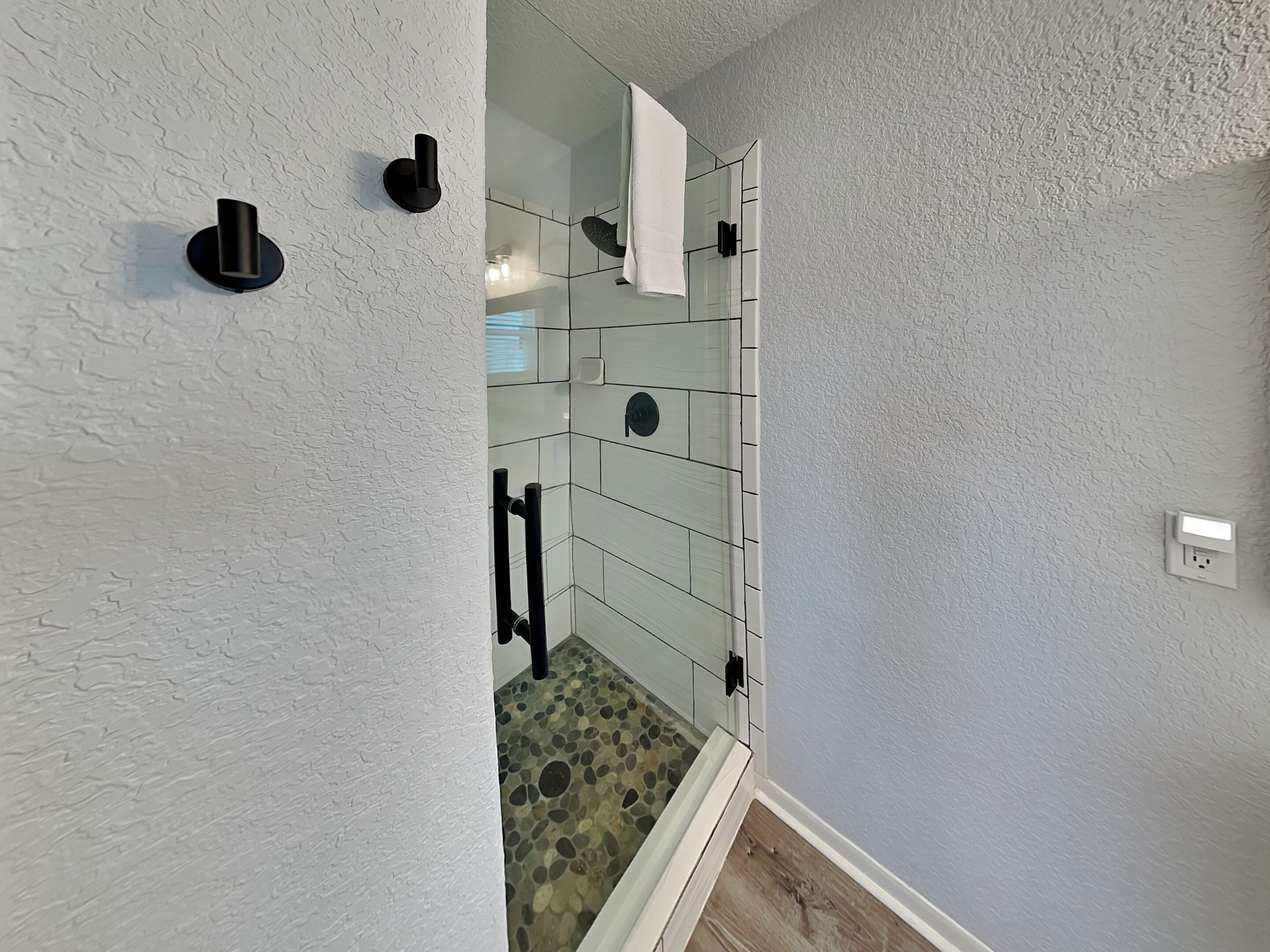 Une salle de bain avec un mur blanc à texture, une zone de douche carrelée et une prise électrique visible.
