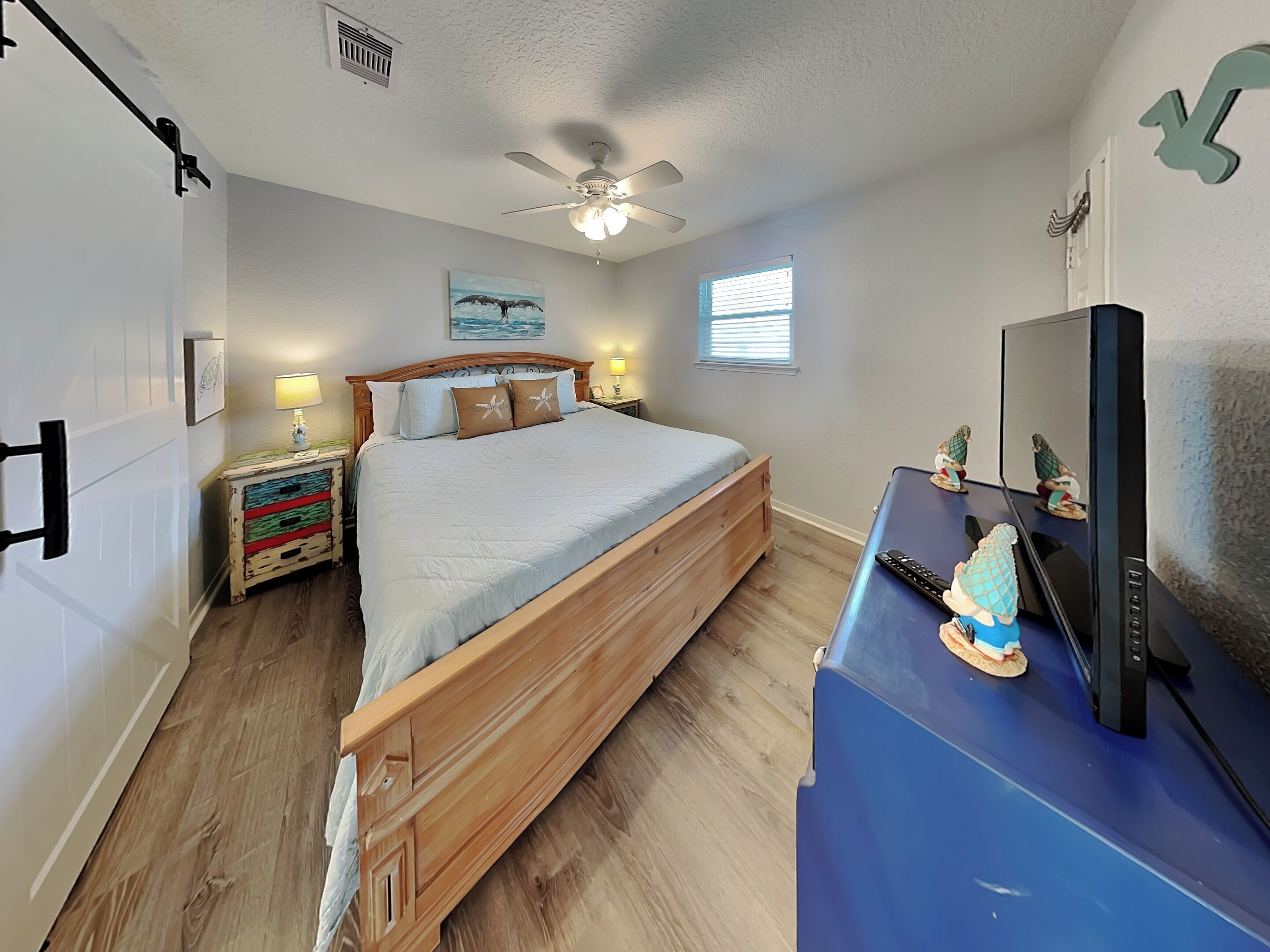 A bedroom with a wooden bed, a ceiling fan, a window with blinds, a dresser with a television, and a small decorative figure.