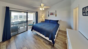 A bedroom with a bed, a ceiling fan, a nightstand, a lamp, a view through a sliding door, and a sign on the wall. - La Buena Vida by Miss Kitty’s (Rockport)