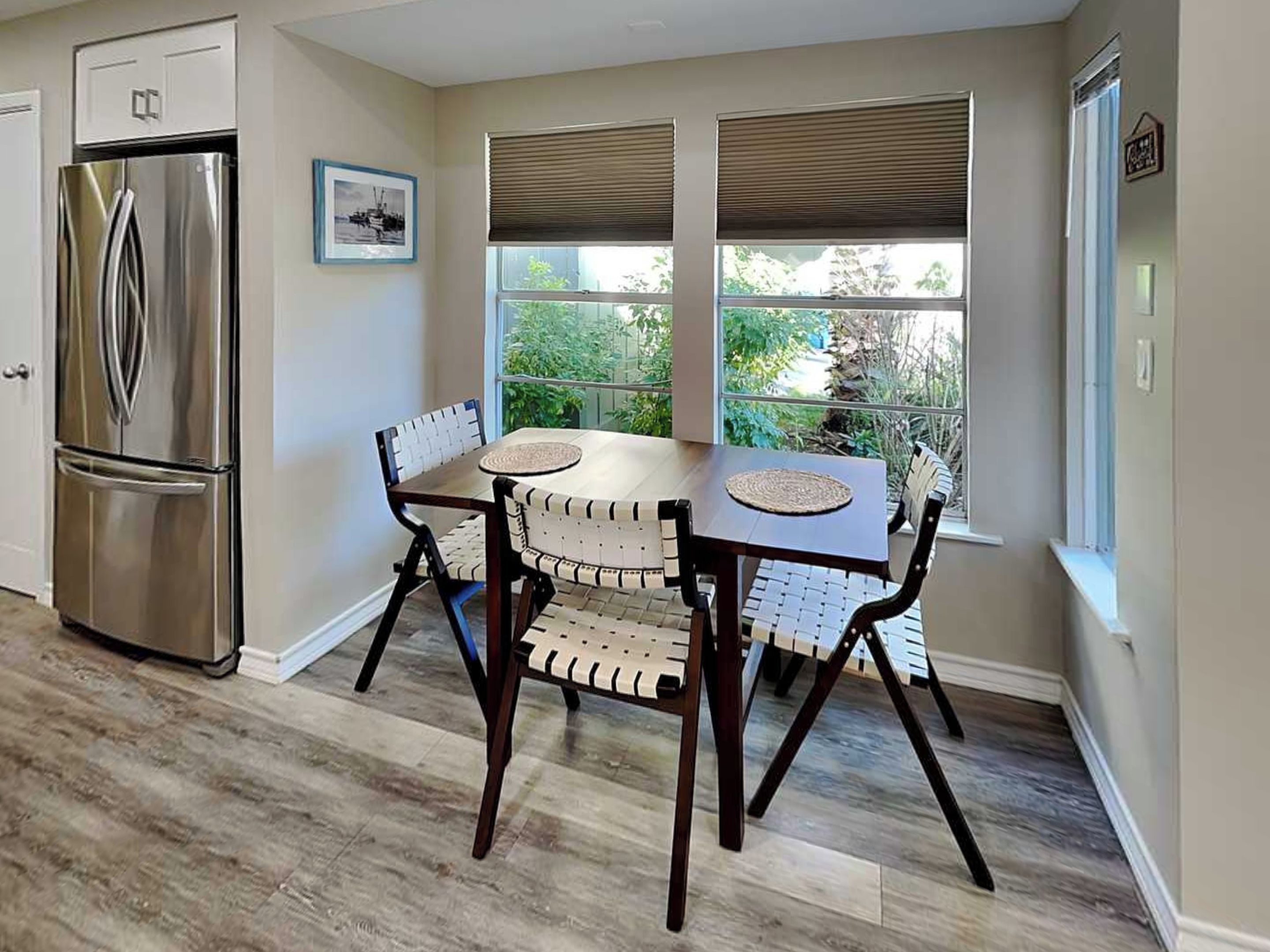 Una cocina con un refrigerador de acero inoxidable, una mesa de comedor con cuatro sillas y una ventana con persianas.