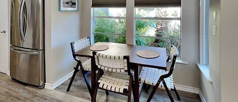 A kitchen with a stainless steel refrigerator, a dining table with four chairs, and a window with blinds.