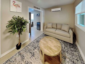 Uma sala de estar com um sofá bege, uma mesa de centro de madeira, uma planta em vaso e um ar-condicionado fixado na parede.
