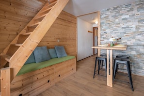 Un interior de madera con una escalera, un sofá verde con cojines, una zona de cocina con taburetes y una pared con diseño de piedra.