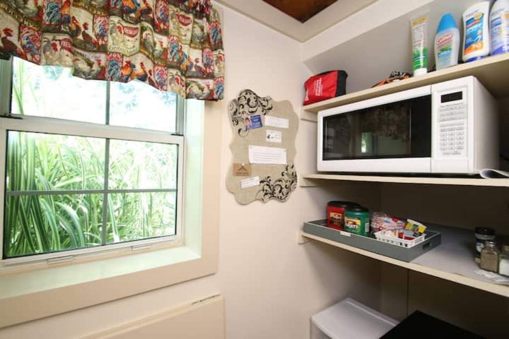 Une cuisine équipée d’un four à micro-ondes, d’étagères remplies de provisions, d’une fenêtre avec des rideaux et d’un tableau d’affichage.