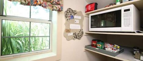 A kitchen with a microwave, shelves with food items, a window with curtains, and a bulletin board.