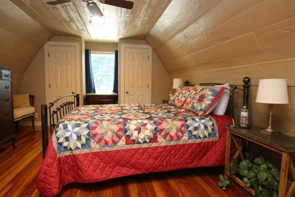 Un dormitorio con edredón acolchado, suelo de madera, ventilador de techo y ventana con cortinas.