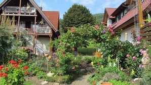 A garden with a variety of plants and flowers, a wooden deck, and a building with a sloped roof. - Herrlich an Wald u. Wiesenrand,ruhige Lage. Erdgeschoss, Terrasse ,fam. mit Kind (Berghaupten)