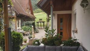A covered patio with a cushioned seating area, a glass-top table, and a view of a garden and buildings in the background. - Herrlich an Wald u. Wiesenrand,ruhige Lage. Erdgeschoss, Terrasse ,fam. mit Kind (Berghaupten)