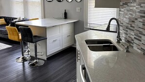 A modern kitchen with a white countertop, a built-in sink, and a backsplash of small tiles. There are two bar stools with black and yellow seats, and a dining table with a yellow cushion. - Lovely 1 bedroom apartment

 (Livingston)