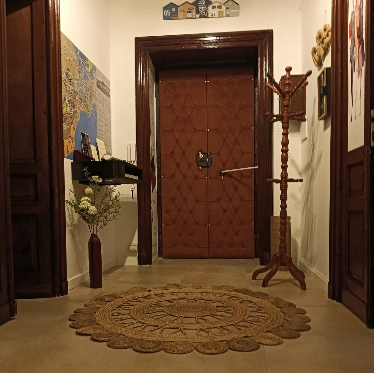 A hallway with a brown door, a wooden coat rack, a vase with flowers, and a decorative rug.
