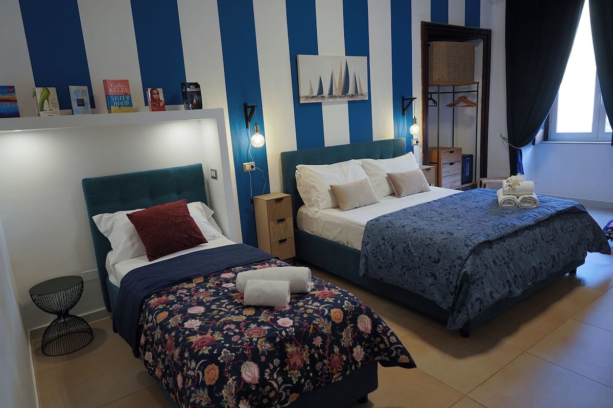 A bedroom with two beds, a bookshelf with books, a wall-mounted artwork, and a window with curtains.