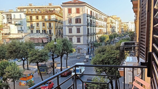 Un balcón con una mesa y sillas, con vistas a una calle con edificios y árboles.
