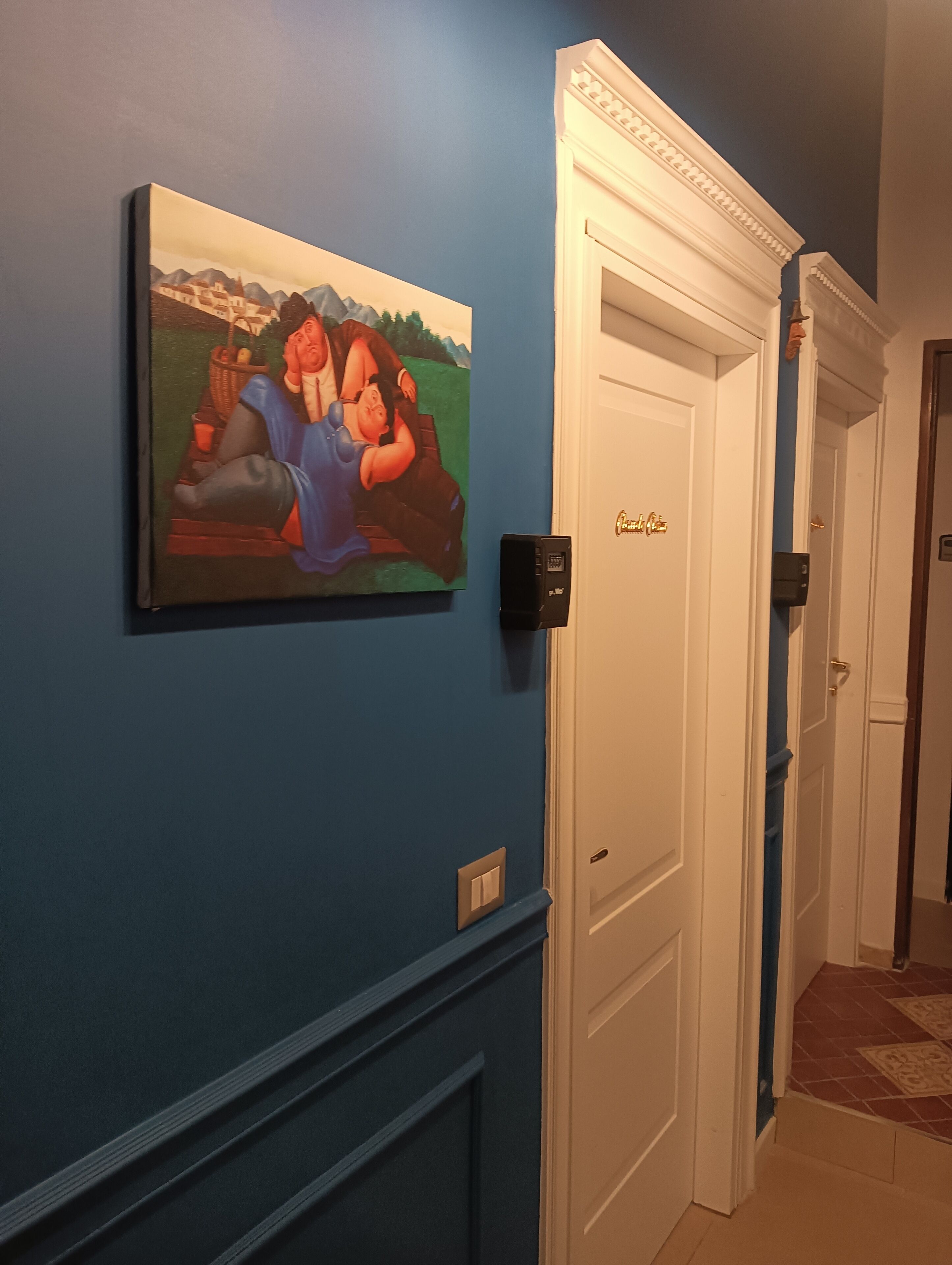 A hallway with a blue wall, a painting, a door with a gold handle, and a light switch.