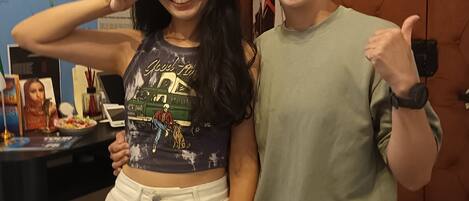 Two people standing in a room with a map and flags on the wall, a wooden door, and a desk with a computer and a photo frame.