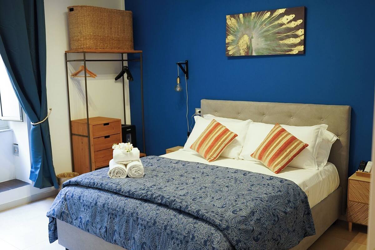 A bedroom with a bed, a bedside table, a hanging light fixture, a wall-mounted air conditioner, and a window with curtains.